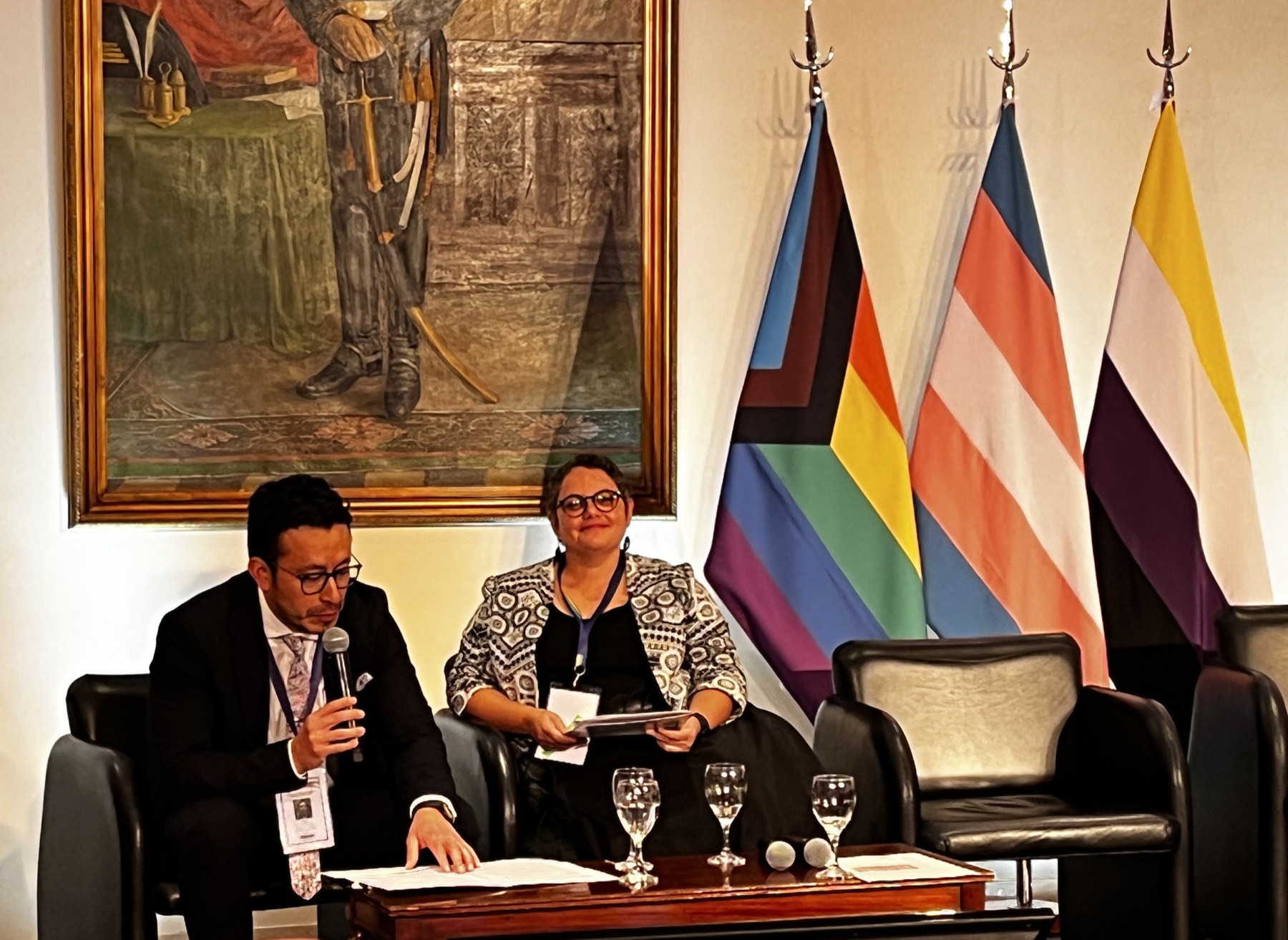 Lana Woolf speaking on a conference panel beside pride, trans and non-binary flags